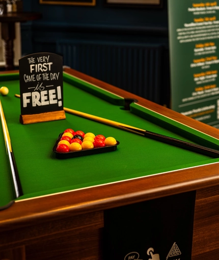 A close up view of the pool table inside the Malvern Tavern, with the balls, cues, and a sign saying "The very first game of the day is free!" on the top.