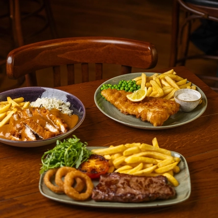 A close up view of a wooden restaurant table laden with a bowl of Katsu Chicken with fries and rice, a plate of Fish & Chips served with peas, lemon, and dipping sauce, and a plate of steak, chips, onions rings, tomato, and salad.