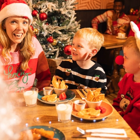 An adult wearing a Santa hat sits with two children at a wooden table in a pub decorated for Christmas. The table is set with glasses of milk, pancakes, strawberries and bowls of crisps, while a snow-covered Christmas tree with red baubles stands nearby. A festive Santa figure and other guests are visible in the background, creating a warm family Christmas breakfast setting.