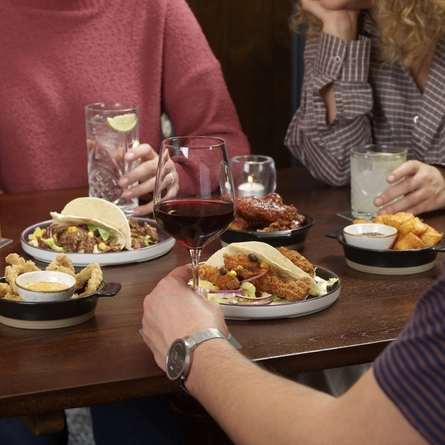 A group of people sitting around a wooden table at a restaurant, sharing several small plates of food including tacos, fried items, and dipping sauces. Drinks are on the table. The individuals are engaged in conversation while eating, with one person holding a piece of food and others seated around the table.