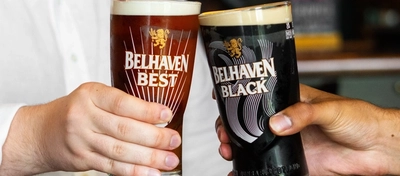 Two people at a wooden table inside a pub, one holding a glass of Belhaven Best ale and the other holding a glass of Belhaven Black stout. They're clinking the glasses together in a cheers motion and two coasters sit on the table in front of them.