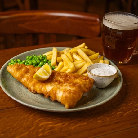 A close up view of Fish & Chips served on a plate with peas, a slice of lemon, and a small pot of dipping sauce. The plate sits on a wooden restaurant table along with a glass of beer.