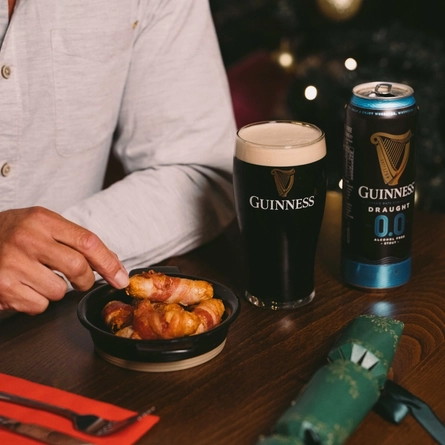 A close up view of a small roasting dish filled with pigs in blankets, sitting on a wooden restaurant table along with a glass of Guinness and a can of alcohol free Guinness. Cutlery, a serviette, and a Christmas cracker also sit on the table; a person at the table is picking up one of the pigs in blankets; and Christmas tree is visible in the background.