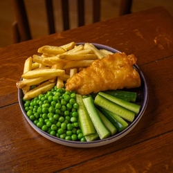 A close up view from above of a children's Fish & Chips meal served on a plate with peas and vegetable sticks. The plate sits on a wooden restaurant table.