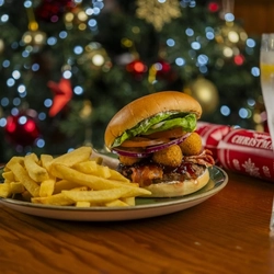 A Camembert burger served on a plate with fries and coleslaw. The plate sits on a wooden table with cutlery, a glass of lemonade, and a Christmas cracker, and a Christmas tree is visible in the background.
