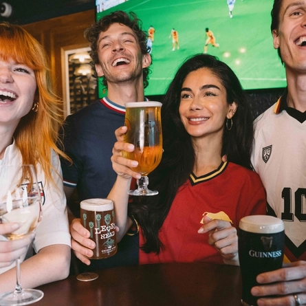 An image showing a group of friends stood at a table watching a football match and enjoying a selection of drinks within the interior seating area at a Greene King venue.