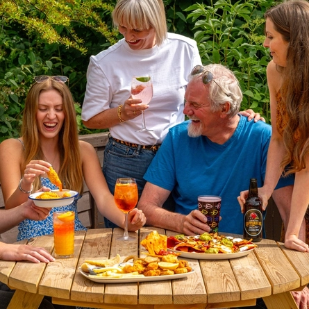 A mixed group of people seated around a wooden table, in a beer garden, enjoying a variety of sharers and drinks.