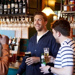 A group of people in a pub bar area, one of them is holding a glass of Guinness, one of them is holding a glass of beer, and another person is holding a bottle of Peroni beer.