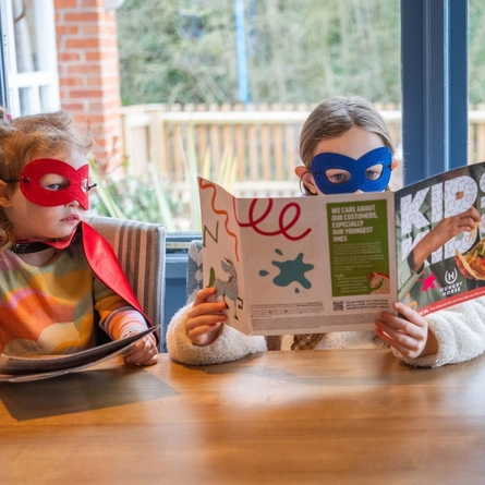 Two children dressed as superheros sat at an indoor wooden table, reading a cgildren's comic, activity book.