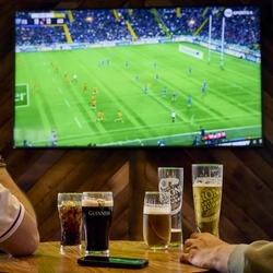 Two people sitting at a table in a pub, watching sports on the TV on the wall in front of them. A round of drinks and a mobile phone sit on the table between them.