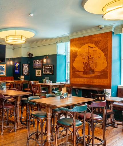 The interior restaurant seating area at the Royal George in Euston; with an upholstered booth seat, a TV on the wall above it, a fireplace, and a large wooden inlay picture of a sailing ship on the wall.