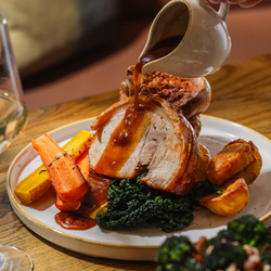 A lifestyle image showing a jug of gravy being poured over a plated Roast Pork Loin dish sat on a table within the interior restaurant seating area at The Four Oaks.