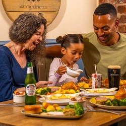 Three people seated around a wooden table in a pub, sharing Sunday roast meals. One person pours gravy from a small jug onto a plate of roast dinner with Yorkshire puddings, vegetables and potatoes. A bottle of alcohol-free beer, a pint of Guinness and a children's juice carton sit among the dishes. The background features blue panelled walls, exposed brickwork and a wooden barrel-style sign reading “The Mill House”.