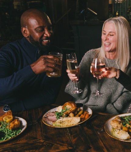 A lifestyle image focusing on 3 customers within a group sat at a table enjoying Mothers Day main dishes and drinks, celebrating with a cheers within the interior restaurant seating area at a Heritage venue.