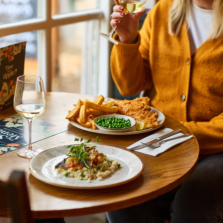 An image of a customer sat within the interior restaurant seating area enjoying a glass of white wine with 2 Main dishes and a glass of white wine sat on the table from the Set menu at a Chef & Brewer venue.