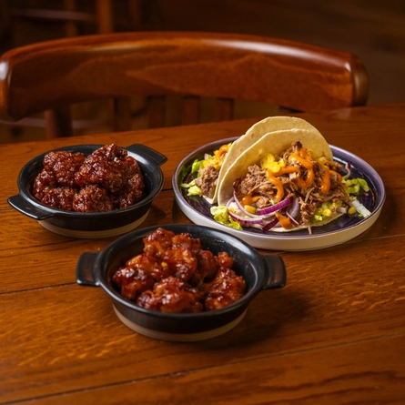 Karage Chicken Wings and Cauliflower Wings served in small roasting dishes, and a plate of tacos, sitting on a wooden restaurant table.