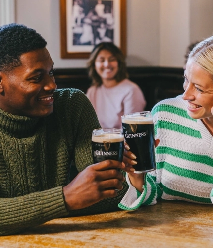 A man and a woman are seated at an indoor wooden table, each holding a pint of Guinness in a Cheers position.
