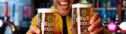 A man standing next to a bar holding two pints of Level Head, smiling at the camera.
