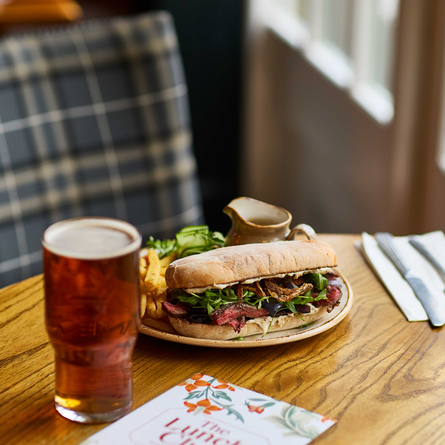 An image focusing on a Steak Ciabatta lunch dish and pint of ale sat on a table within the interior restaurant seating area at a Chef & Brewer venue.