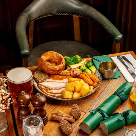 A plated turkey dinner on a festive decorated wooden table with a tankard of beer, a glass and bottle of sparkling wine and a stemmed glass containing a spirit cocktail.