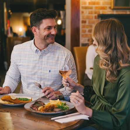 Two people sitting side by side at a restaurant table, one of them holding a glass of wine. Two plates of food and a glass of Hazy Day IPA sit on the table in front of them.