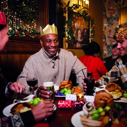 A group of people are gathered around a table enjoying a festive meal. They are wearing paper crowns, and the table is set with plates of food. The background is decorated with Christmas decorations.