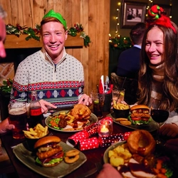A group of people are gathered around a table enjoying a festive meal. They are wearing paper crowns, and the table is set with plates of food. The background is decorated with Christmas decorations.