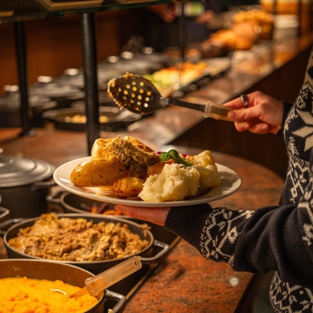 A person standing at a carvery counter holding a plate which is loaded with Yorkshire pudding, new potatoes, roast potatoes, mashed potato, and vegetables, while using a large spoon to put stuffing onto the plate.