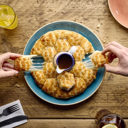 A bird's eye view of a Garlic Bread Tear & Share served on a plate with a jug of sauce in the centre; and two hands tearing off sections of the bread to reveal stretchy melted cheese inside. Cutlery, serviettes, two other plates, and two glasses of soft drink also sit on the table.