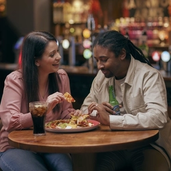 A man and woman sitting at a table with a bottle of Heineken and a glass of Coca Cola, sharing a plate of nachos.