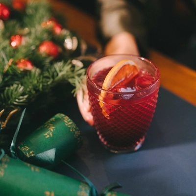 A hand resting on a festive decorated table is holding a cocktail type drink in a lowball glass.