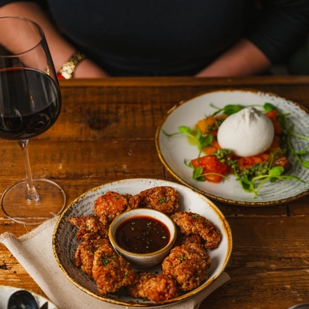 Pieces of Karage Chicken served in a bowl with a small pot of dipping sauce. The bowl sits on a wooden table along with two other plates of food, a glass of red wine, and a tea light, and a napkin is tucked under the bowl.