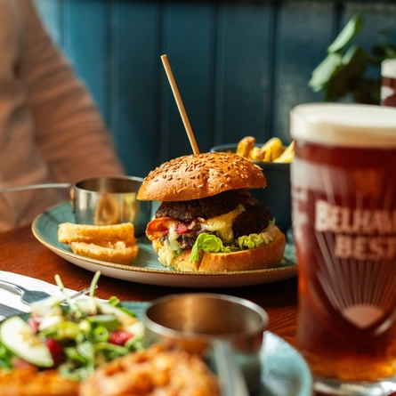 A Balmoral Burger served on a plate with onion rings, a small bowl of fries and pots of sauce. The plate sits on a wooden restaurant table with a glass of Belhaven Best ale, cutlery, a napkin, and another plate of food.