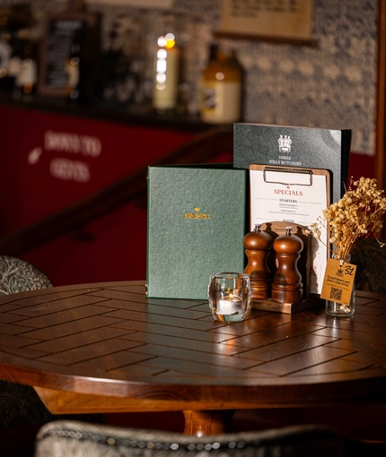 A close up of the interior restaurant table showing a display that consists of menus, salt and pepper shakers, candles and vases with decorative flowers at the Three Jolly Butchers.