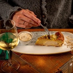 A person is sitting at a wooden table, holding a fork and cutting into a slice of cake on an oval plate.