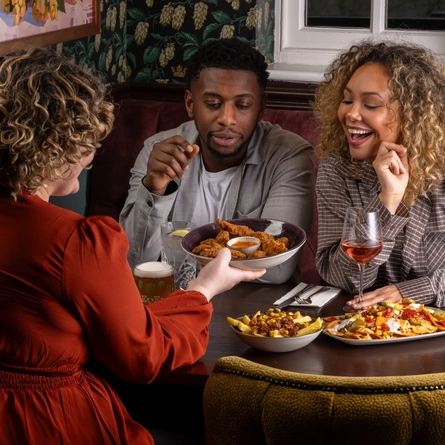 Three people sit around a wooden table in a restaurant booth. One person seated on the far side of the table is holding a piece of fried food toward the person sitting beside them. A third person, is seated facing the pair and reaching forward with a large bowl filled with shared appetizers. Other shared dishes are spread across the table.