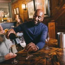 A lifestyle image focusing on a customer sat at a table with friends pouring out glasses of sparkling white wine to celebrate Mothers Day within the interior seating area at a Heritage venue.