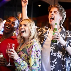 A group of people inside a pub bar area, laughing, singing and dancing. One holds a bottle of beer, one holds a glass of wine, one holds a glass of beer and another holds a cocktail in a wine glass.