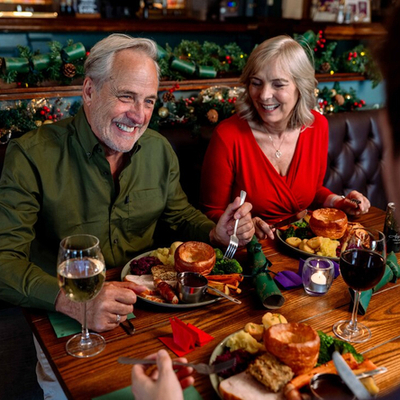 An image focusing 2 people sat within a group at a table within the interior restaurant seating area enjoying drinks and dishes from the Christmas menu at a Community Social venue.