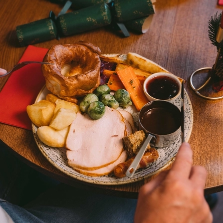 A close up view of a plate of Christmas dinner including Yorkshire pudding, turkey, roast potatoes, vegetables, Brussels sprouts, and a pot of gravy; sitting on a wooden restaurant table. A glass of white wine, two green Christmas crackers, a red serviette, and a red taper candle also sit on the table, and a person at the table is holding their knife and fork above the plate.