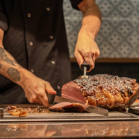 A close up view of a joint of roast beef sitting on a tray on a carvery counter while a member of staff carves it into slices.