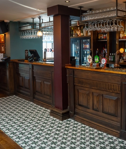 The wood panelled bar at The Ship; with a patterned tile floor in front of the bar, golden drinks trays on the counter, and glasses hanging in racks overhead.