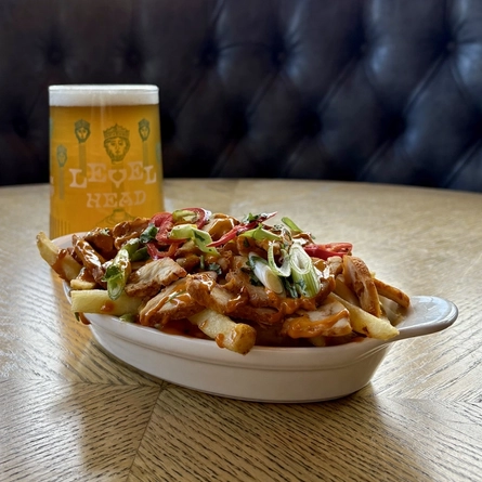 A roasting dish containing Loaded Fries, with a Pint of Level Head, on an indoor table.