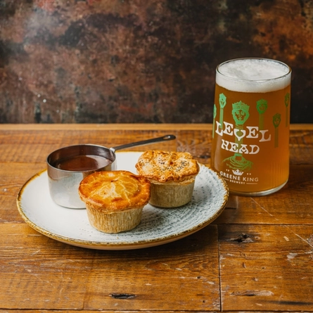 An advertising image showing 2 pies and a jug of gravy on a plate sat on a wooden table next to a pint of Level Head to advertise a Pie & Pint offer available at Urban Social & Community Social venues.