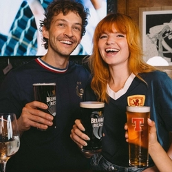 An image focusing on 2 people within a group of friends stood at a table watching a football match and enjoying a selection of drinks within the interior seating area at a Greene King venue.