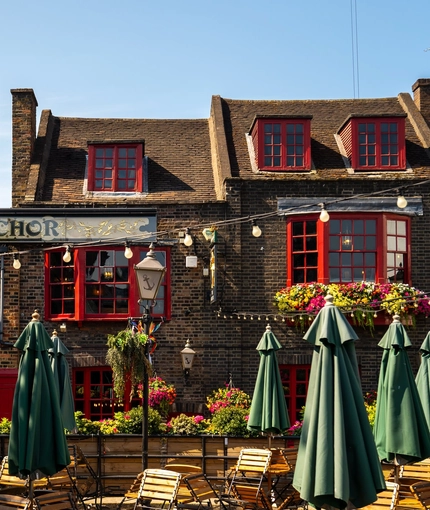 The exterior facade, signage, and beer garden seating area of The Anchor Bankside in Southwark; with shade umbrellas on some of the tables; string lights overhead; and wooden planter boxes filled with flowers.