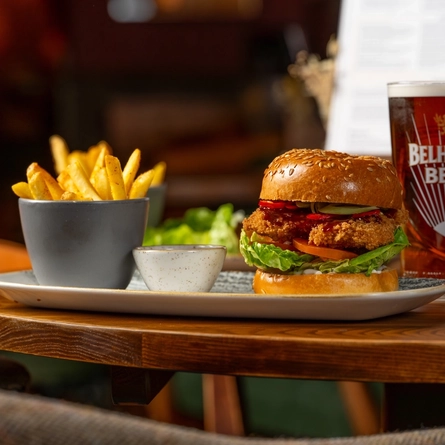 A close up view of a burger, a small bowl of fries, and a pot of sauce served on a plate which sits on a wooden restaurant table along with a glass of Belhaven Best ale and other plates of food.