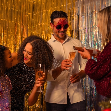Four people gather in front of silver and gold metallic party backdrops during a festive New Year’s celebration. Three adults hold champagne flutes while one person wearing novelty red “Happy New Year” glasses stands in the centre. A child wearing a sequinned outfit stands nearby. A decorated Christmas tree with red and gold baubles and warm fairy lights is visible in the foreground, adding seasonal detail to the party setting.