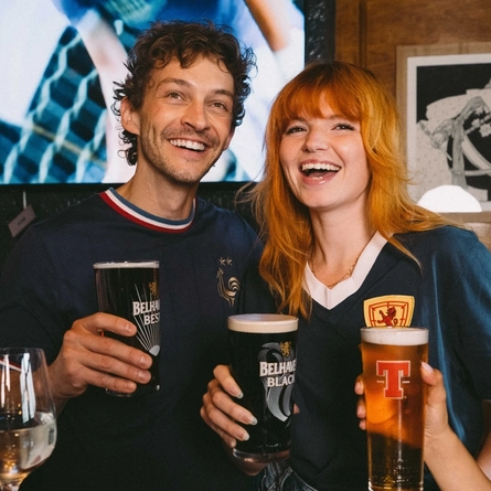 An image focusing on 2 people within a group of friends stood at a table watching a football match and enjoying a selection of drinks within the interior seating area at a Greene King venue.