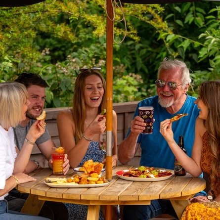 A mixed group of people seated around a wooden table, under an umbrella in a beer garden, enjoying a variety of sharers and drinks.